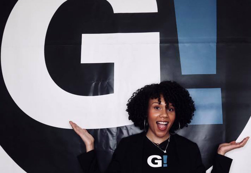 Smiling young black woman against a backdrop with the Girls Be Heard logo